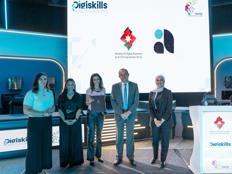 Five individuals, three women and two men, stand on a stage in front of a large screen displaying logos including "Digiskills," "Ministry of Digital Economy and Entrepreneurship," and "Youth Technology Hub." One woman in the center holds a document. The setting appears to be a formal event, likely a signing ceremony or grant award. This image represents Avertra winning a grant from the Ministry of Digital Economy and Entrepreneurship (MoDEE) to upskill Jordan's youth in ICT